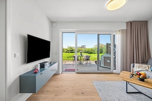 Ein modernes Wohnzimmer mit Flachbildfernseher, einem grauen Unterhaltungsschrank, Holzboden, einer Glasschiebetür zu einer Außenpatio und einem Essbereich mit Tisch und Stühlen.