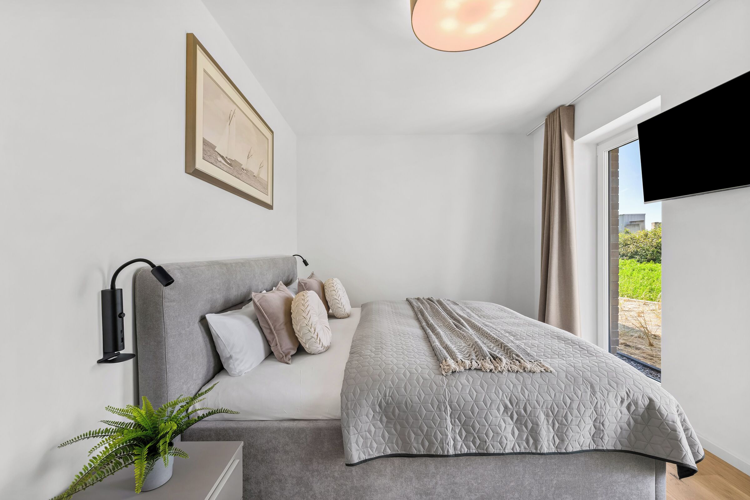 A modern bedroom with a bed, a bedside lamp, a framed picture on the wall, and a window with curtains.