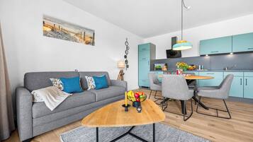 Een moderne woonkamer met een grijze bank, een houten salontafel, en een eethoek met grijze stoelen en een blauwgroene keuken op de achtergrond.