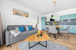 Ein modernes Wohnzimmer mit einem grauen Sofa, einem Holztisch und einem Essbereich mit grauen Stühlen sowie einer türkisblauen Küche im Hintergrund.