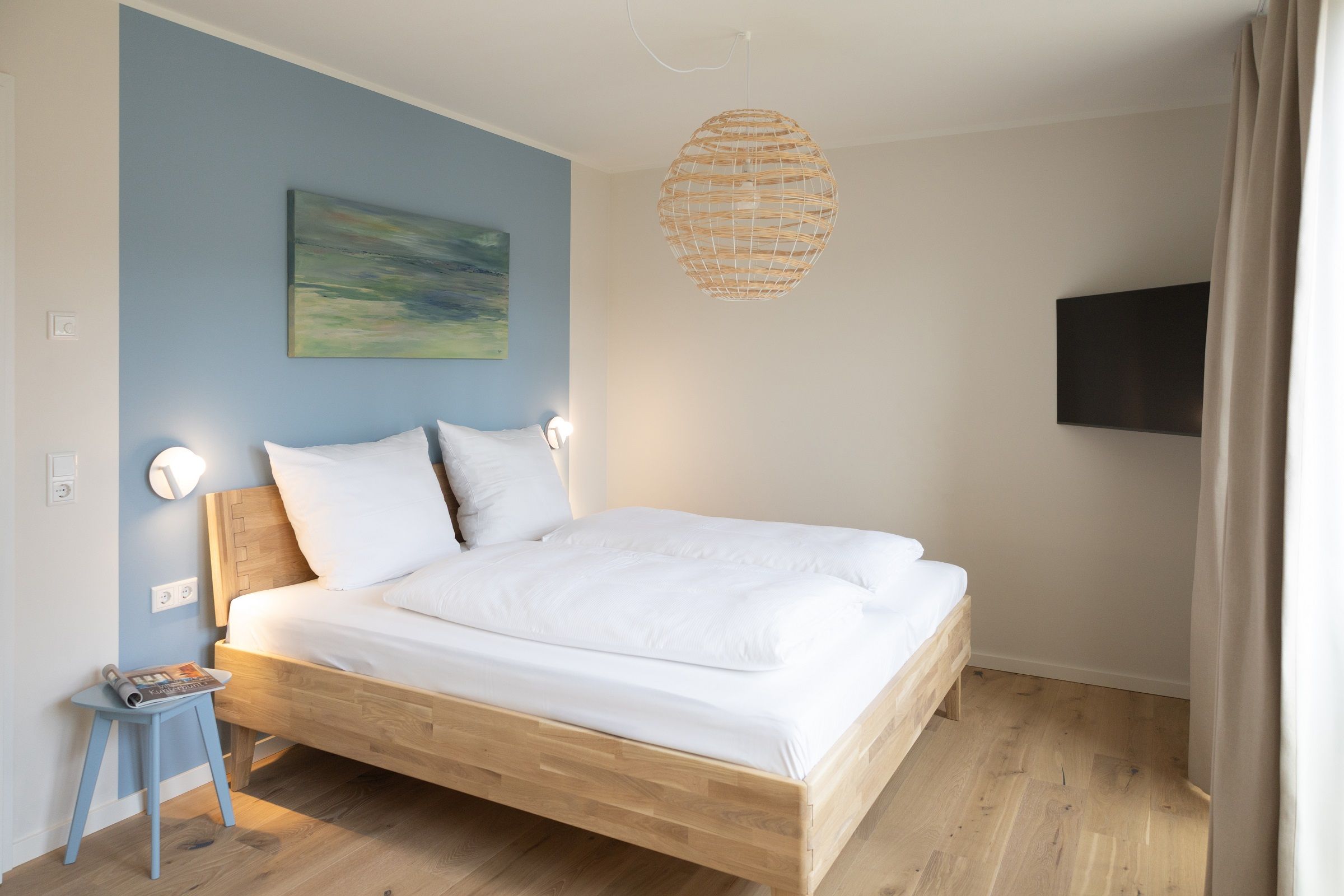A bedroom with a wooden bed, a blue wall, a painting, a woven light fixture, and a television on the wall.