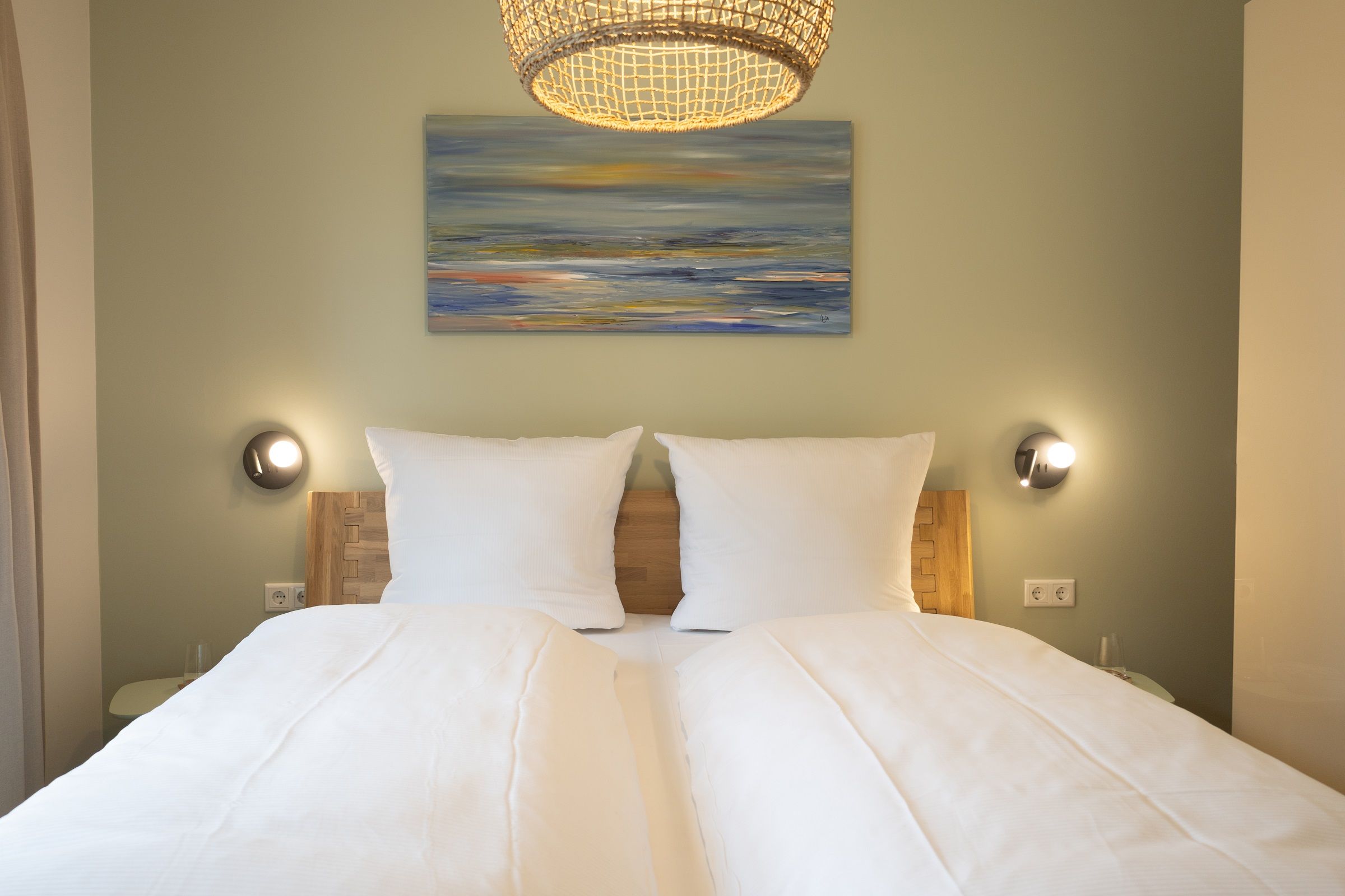 A double bed with white linens, two wall-mounted lamps, and a painting above the bed.