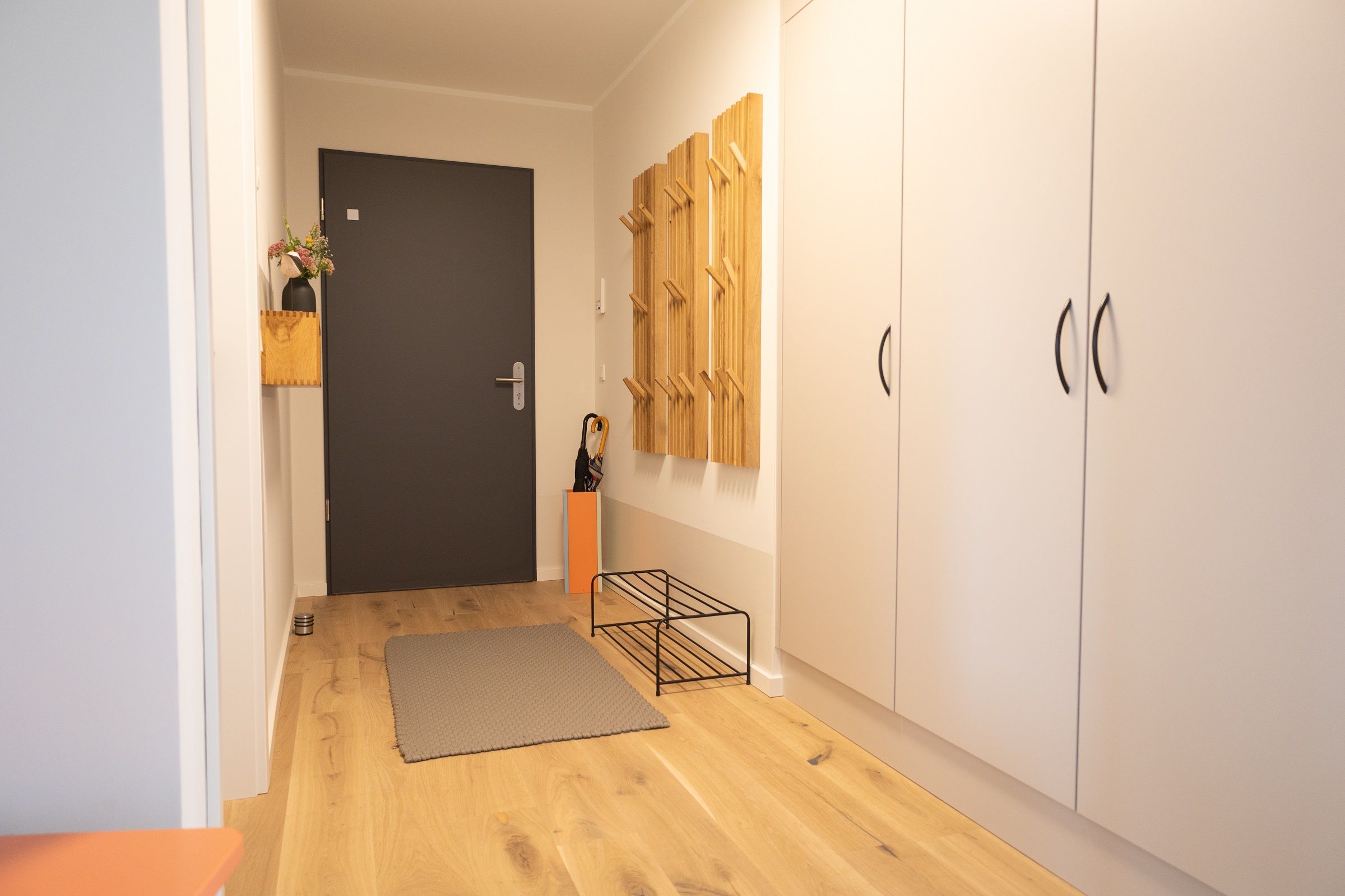 A hallway with a wooden floor, a grey rug, a black door, and a white wardrobe with wooden hooks.