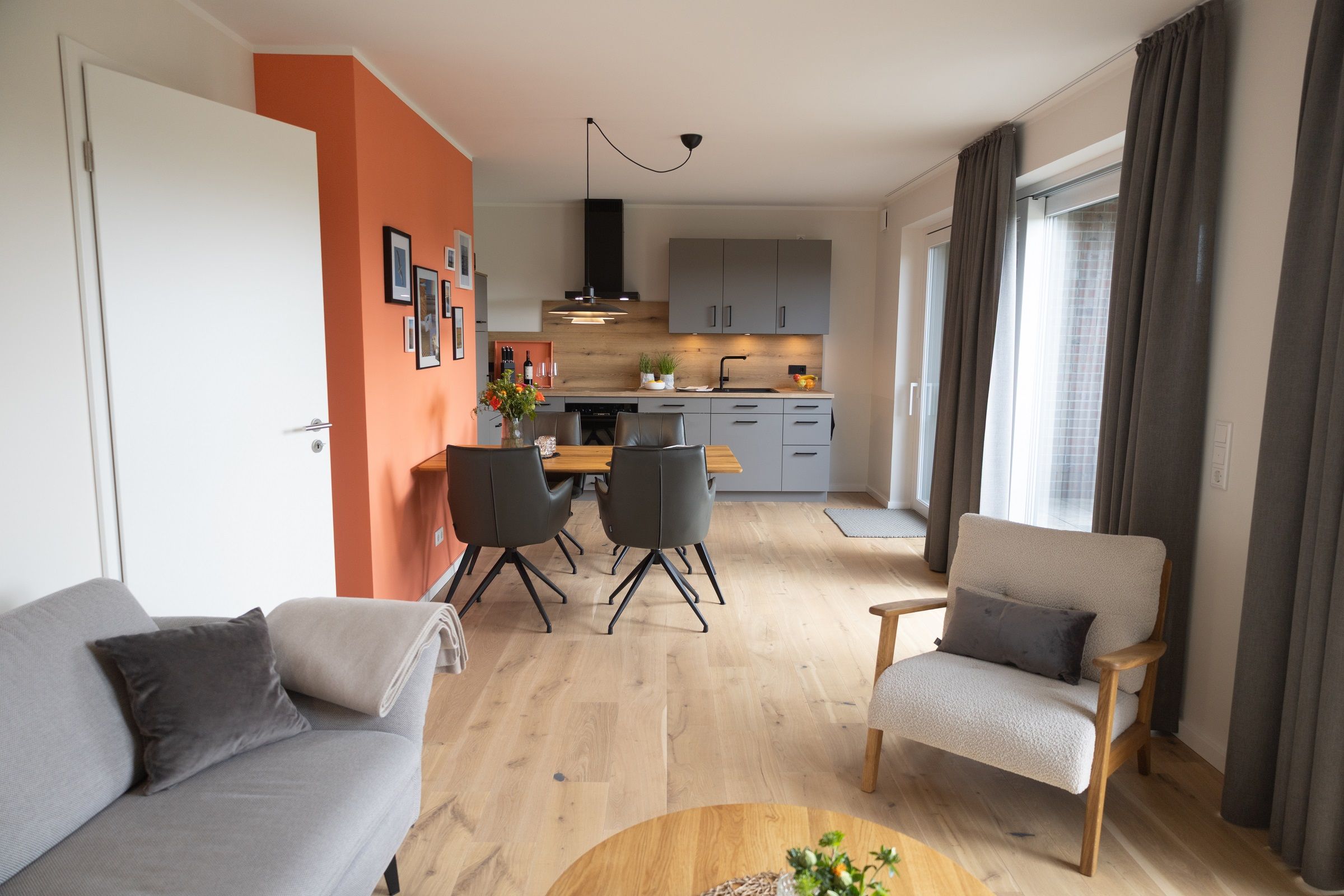 En moderne stue med sofa, lænestol og sofabord. Der er et spiseområde med bord og stole, og et køkken i baggrunden.
