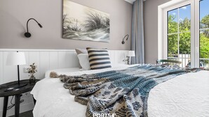 Ett modernt sovrum med en stor säng, en tavla, en lampa och utsikt över grönskan utanför.