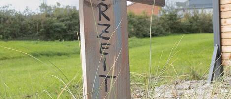 Un letrero de madera con la palabra 'MERZEIT' y dos pájaros en la parte superior, sobre un campo de hierba con un edificio al fondo.