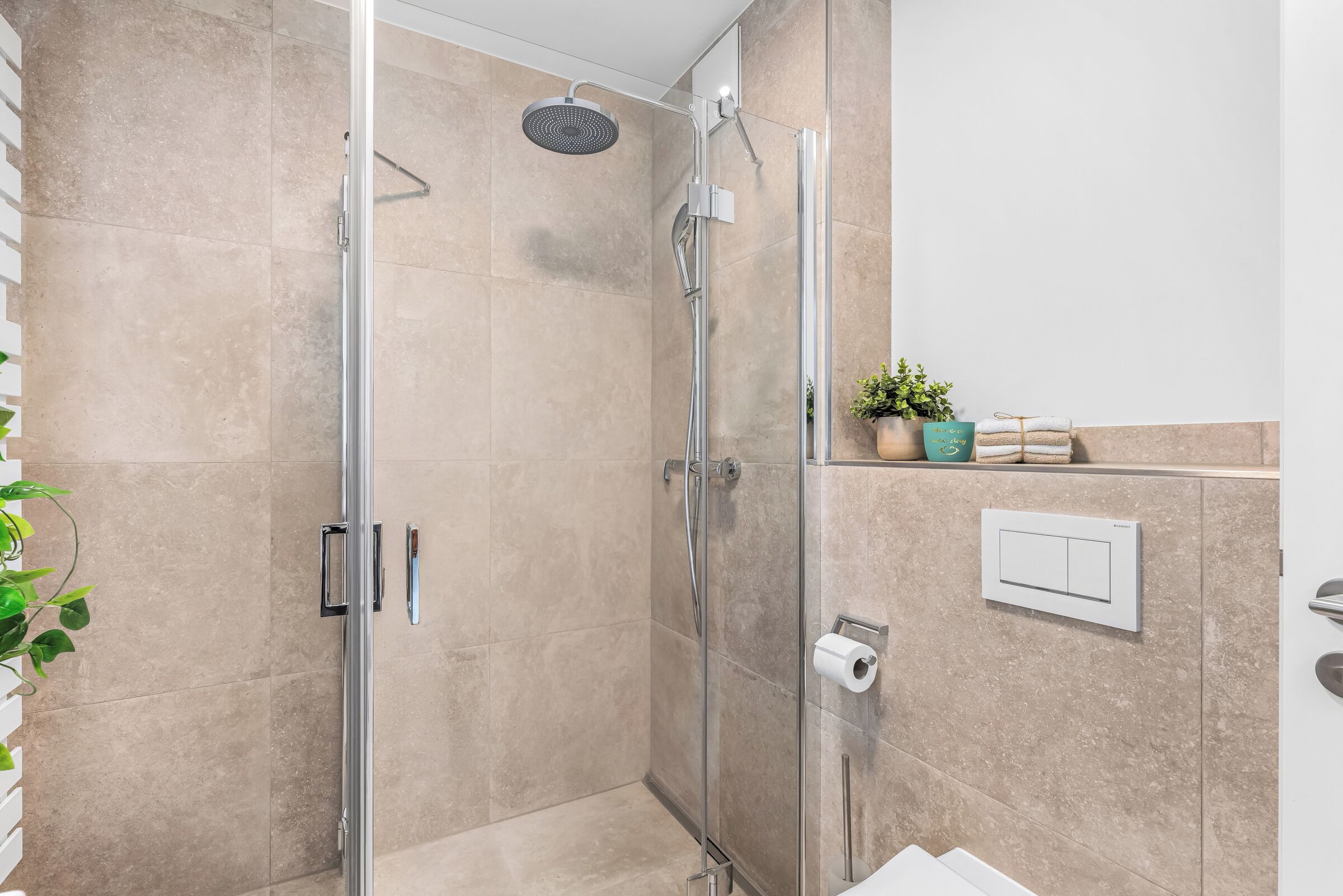 A modern bathroom with a glass-enclosed shower, a towel rack with folded towels, a potted plant, and a toilet paper holder.