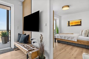 Una sala de estar moderna con un televisor de pantalla plana montado en la pared, un banco de madera con cojines y una cama con colcha blanca.