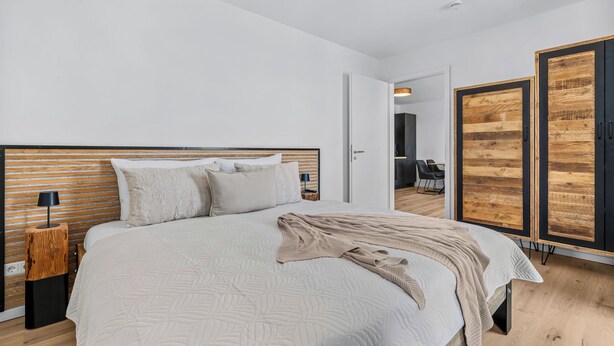 Ein modernes Schlafzimmer mit einem großen Bett, einer Nachttischlampe und einem Schrank mit Holzoptik.