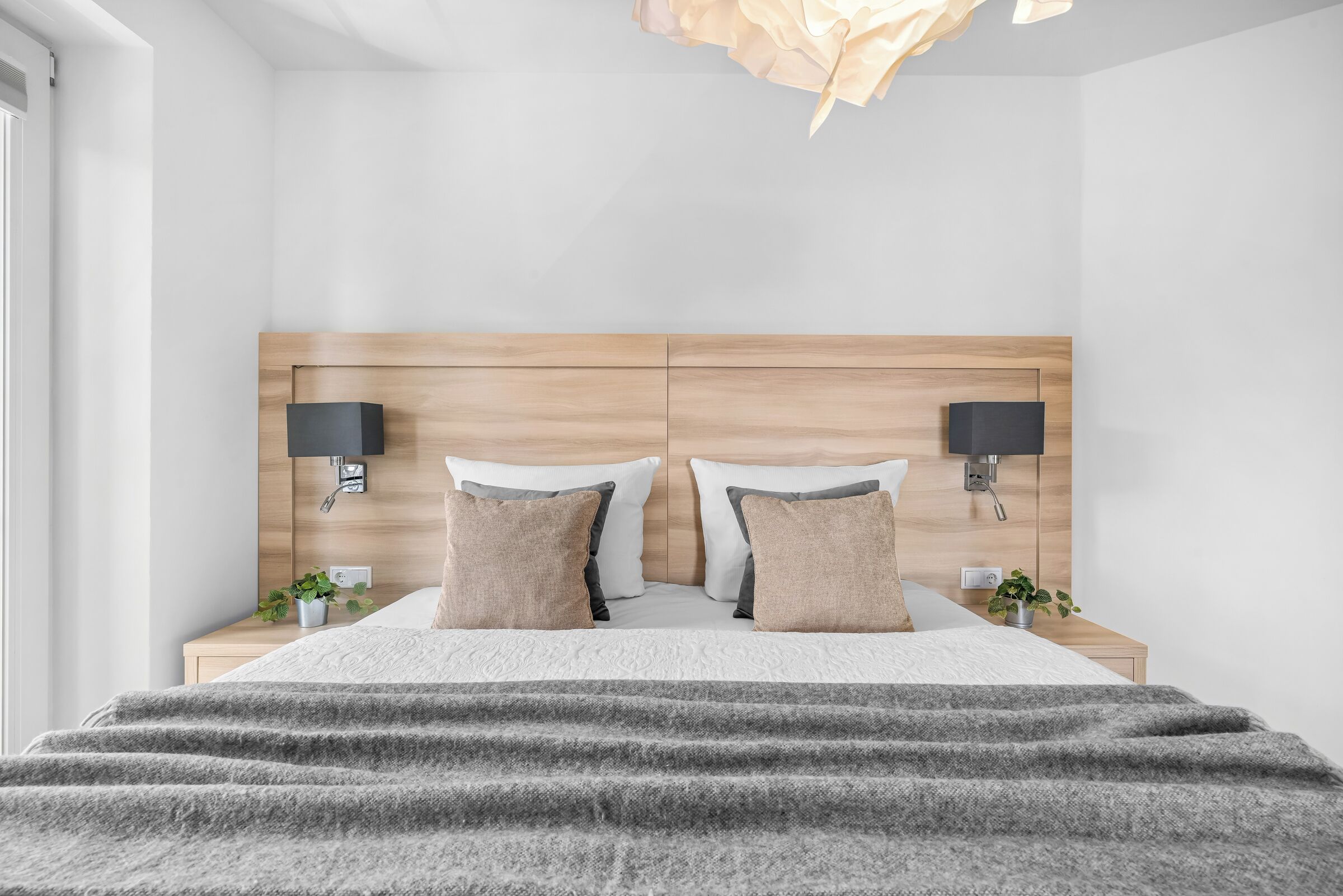 A modern bedroom with a wooden headboard, two bedside tables, a bed with a grey blanket, two wall-mounted lamps, and a small plant on each side.
