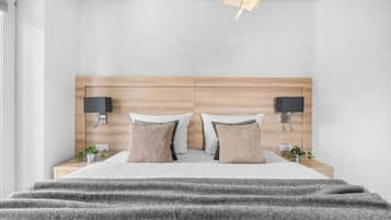 A modern bedroom with a wooden headboard, two bedside tables, a bed with a grey blanket, two wall-mounted lamps, and a small plant on each side.