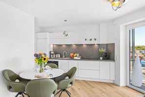 A modern kitchen with white cabinetry, a dining area with green chairs, and a view through a large window. - The View A9 Auster – Sea view, shared sauna, elevator, parking space (Dagebüll)