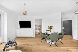A modern living room with a flat-screen TV, a dining area with green chairs, and a kitchen in the background. - The View A9 Auster – Sea view, shared sauna, elevator, parking space (Dagebüll)
