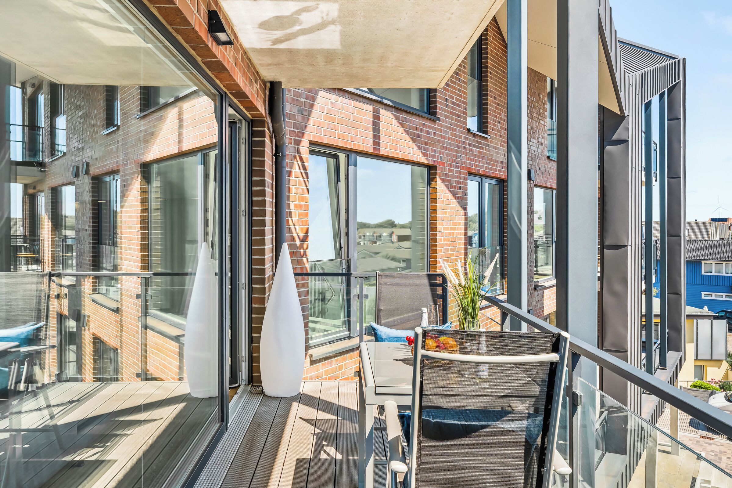 A modern balcony with a glass railing, a white vase, and a chair with a basket of fruit.