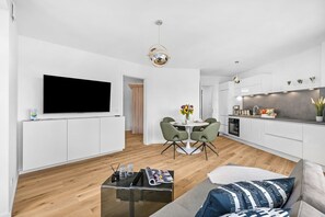A modern living room with a flat-screen TV, a dining area with a round table and chairs, and a kitchen with white cabinets and a built-in oven. - The View A9 Auster – Sea view, shared sauna, elevator, parking space (Dagebüll)