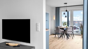 Ein modernes Wohnzimmer mit Fernseher, einer dunklen Ablage, einem Essbereich mit Tisch und Stühlen sowie einem großen Fenster mit Blick auf Gebäude.