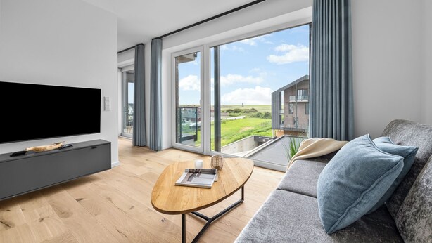 Ein modernes Wohnzimmer mit Flachbildfernseher, Couchtisch aus Holz, einer Couch mit Kissen und großen Fenstern mit Blick auf ein Gebäude und Grünflächen.
