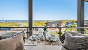 Ein Balkon mit Esstisch, der auf eine Küstenlandschaft mit Windkraftanlagen und Wohnhäusern blickt.
