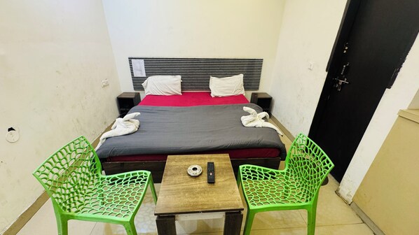 A small, simple room with a bed, two green chairs, a small table, and a single remote control. - Starkey Inn (Gurugram)