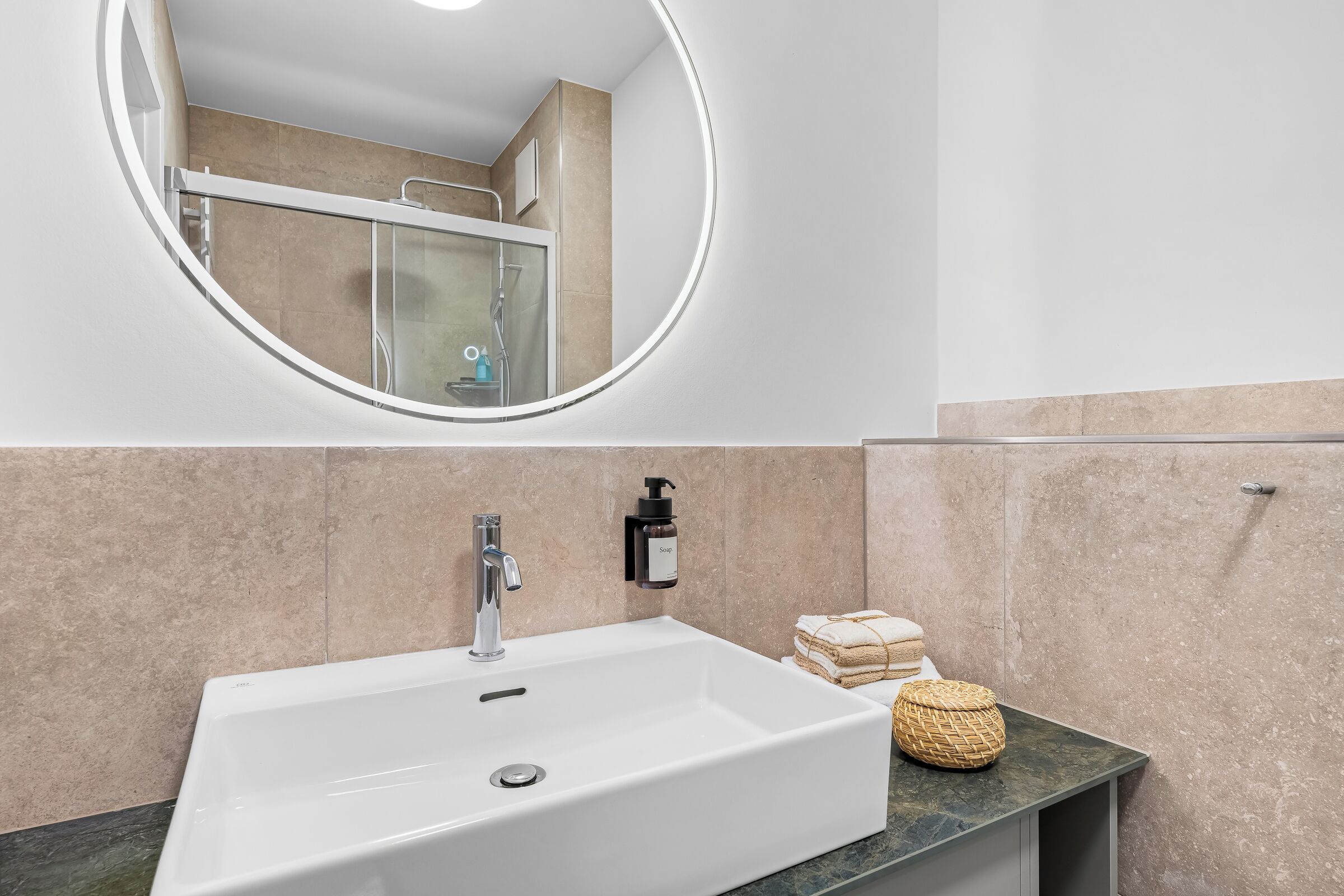 Ein Badezimmer mit einem weißen Waschbecken, einem runden Spiegel und einer Glasdusche.
