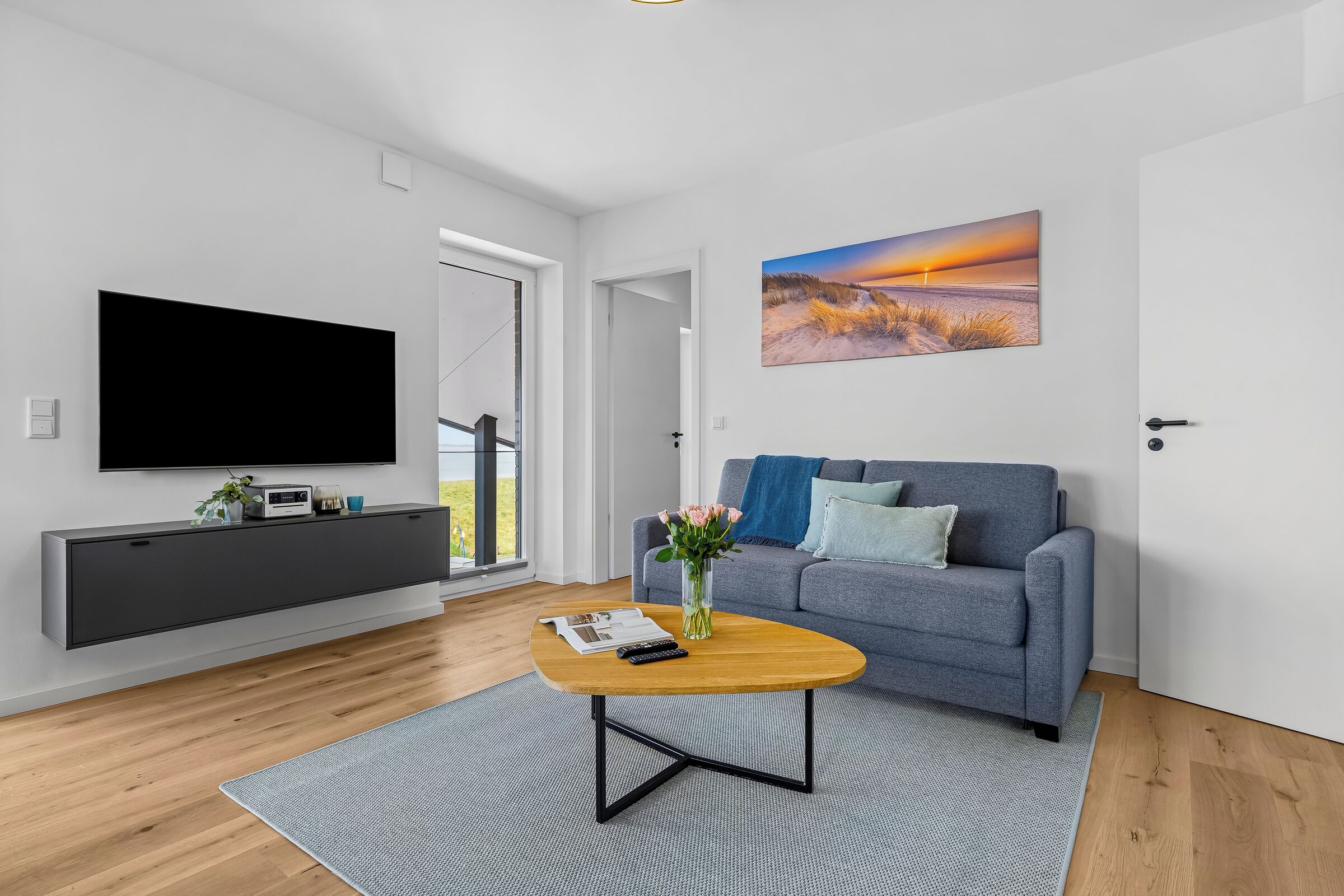 A modern living room with a flat-screen TV, grey sofa, and a painting of a sunset.
