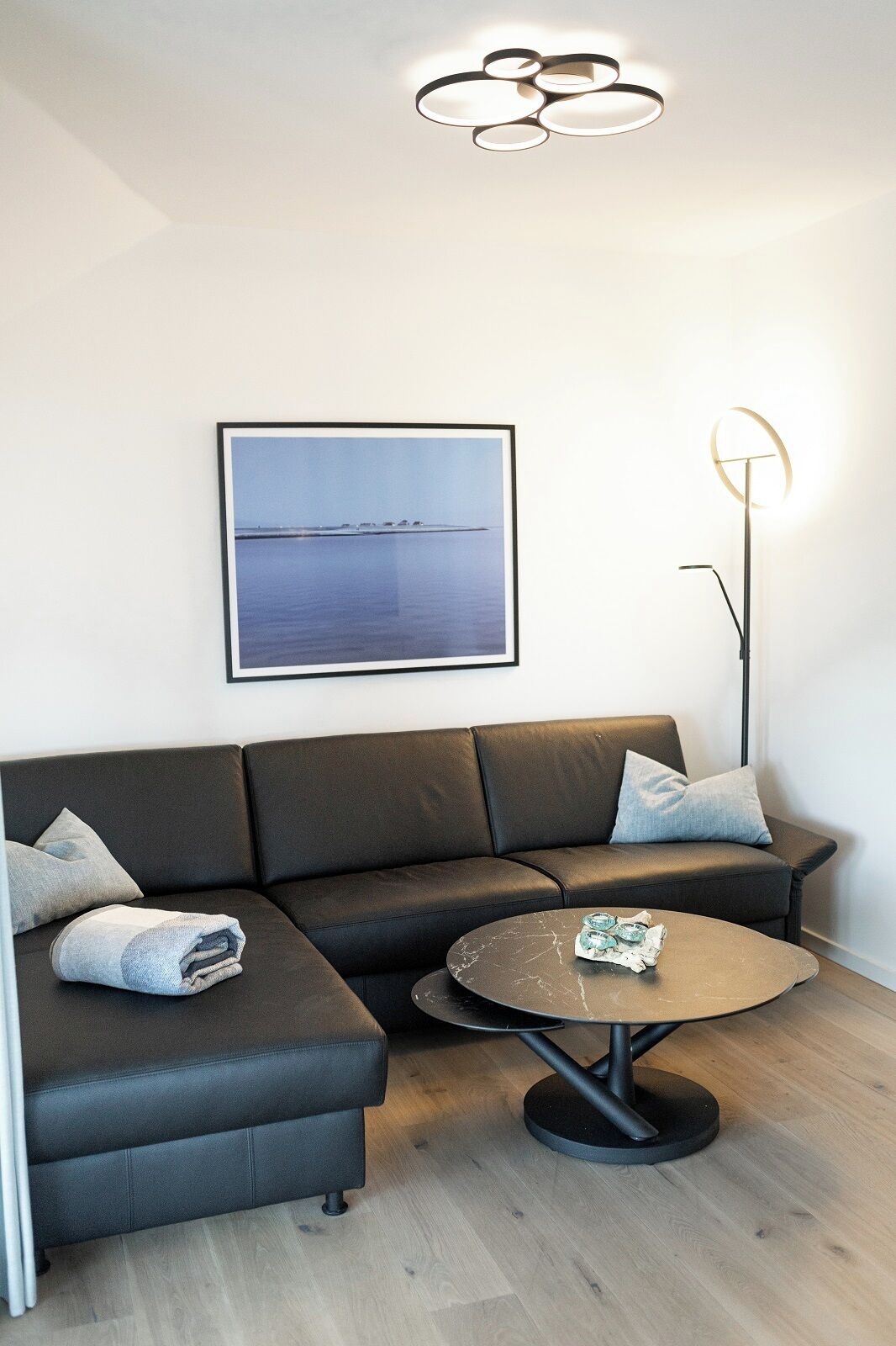 Ein modernes Wohnzimmer mit einem Ecksofa, einem runden Couchtisch, einer Stehlampe und einem gerahmten Landschaftsbild.
