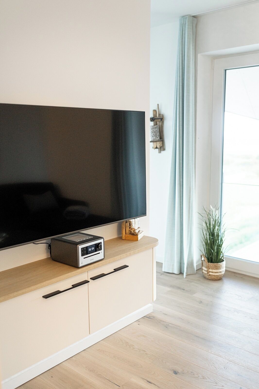 A modern living room with a flat-screen TV mounted on a light-colored cabinet, a small speaker, and a potted plant by a window with light curtains.