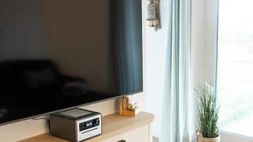 A modern living room with a flat-screen TV mounted on a light-colored cabinet, a small speaker, and a potted plant by a window with light curtains.