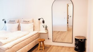 Un dormitorio con cama, almohadas, mesita de noche, espejo, taburete y cubo de basura.