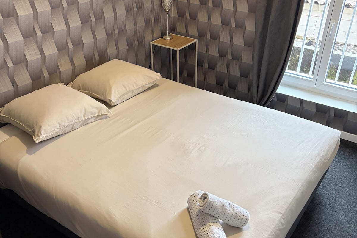 Ein ordentlich bezogenes Bett mit Kissen und einem gerollten Handtuch, ein kleiner Nachttisch mit Lampe und ein Fenster mit Vorhängen.
