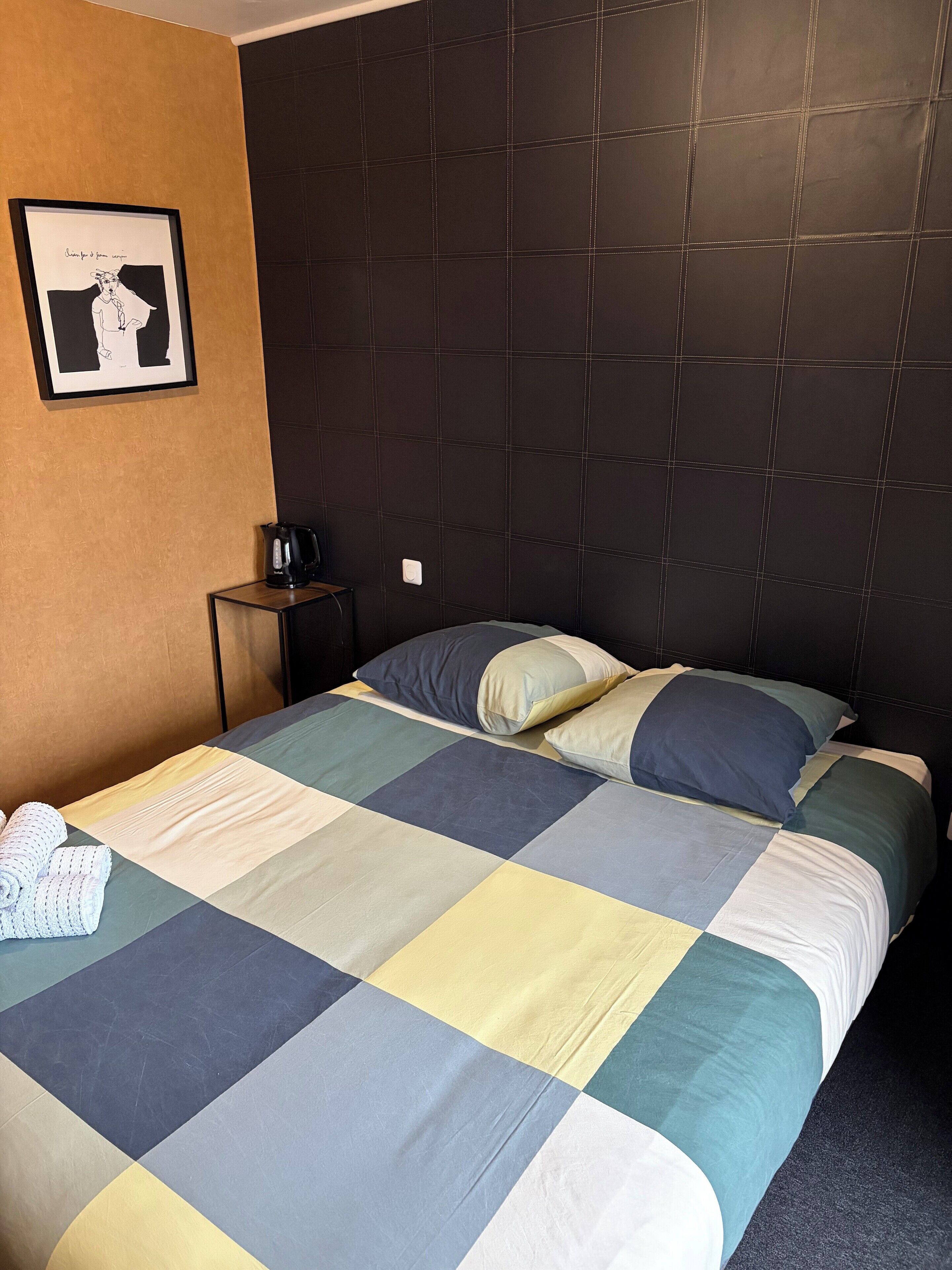 A hotel room with a large bed, a headboard, a framed picture on the wall, a small table with a kettle, and a folded towel on the bed.