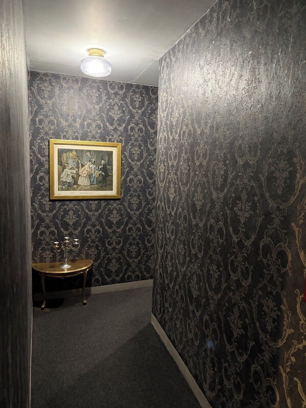A narrow hallway with dark patterned wallpaper, a framed picture on the wall, and a small table with a candelabra. - Hôtel Le Seysses (Seysses)