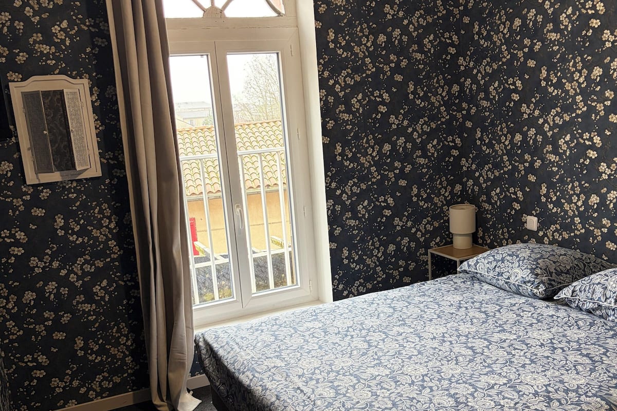 Ein Schlafzimmer mit Bett, Nachttisch, Lampe und einem fenster mit Vorhängen.
