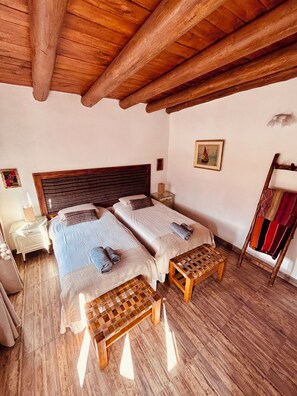 A room with two beds, wooden benches, a small table, a nightstand, and a wall-mounted rack. - La Nochera Killa Cabins (Tilcara)