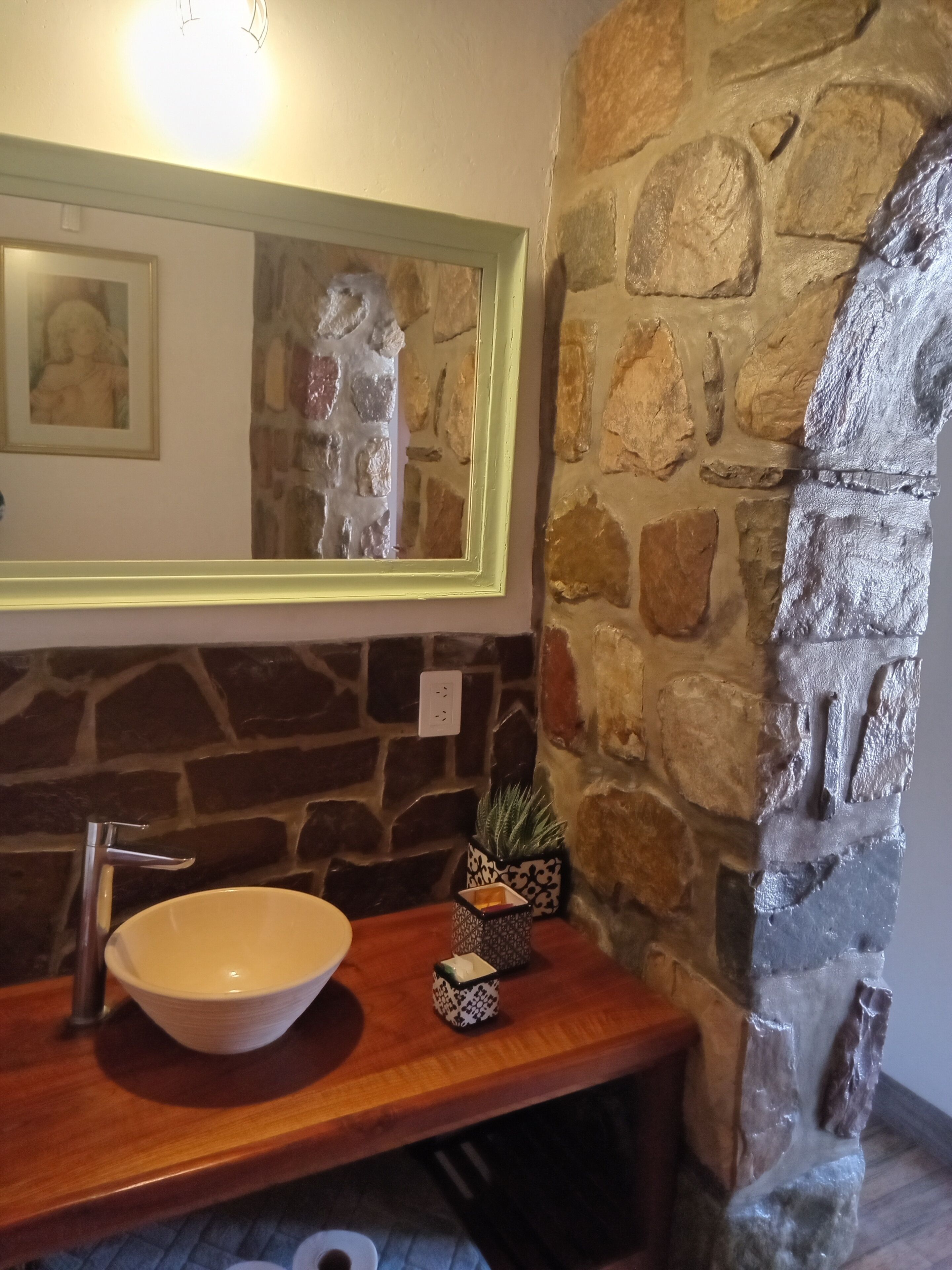 Un baño con pared de piedra, tocador de madera, lavabo blanco, espejo y una pequeña planta en maceta.