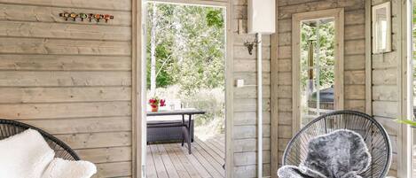 A wooden cabin interior with a door leading to an outdoor area, a clock on the wall, and two chairs with fluffy cushions.