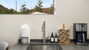 A kitchen with a stainless steel sink, a paper towel dispenser, a coffee maker, and a window with a view of trees and hills. - NEW Cimaron Butte Studio Laundry Kitchenette (Sedona)
