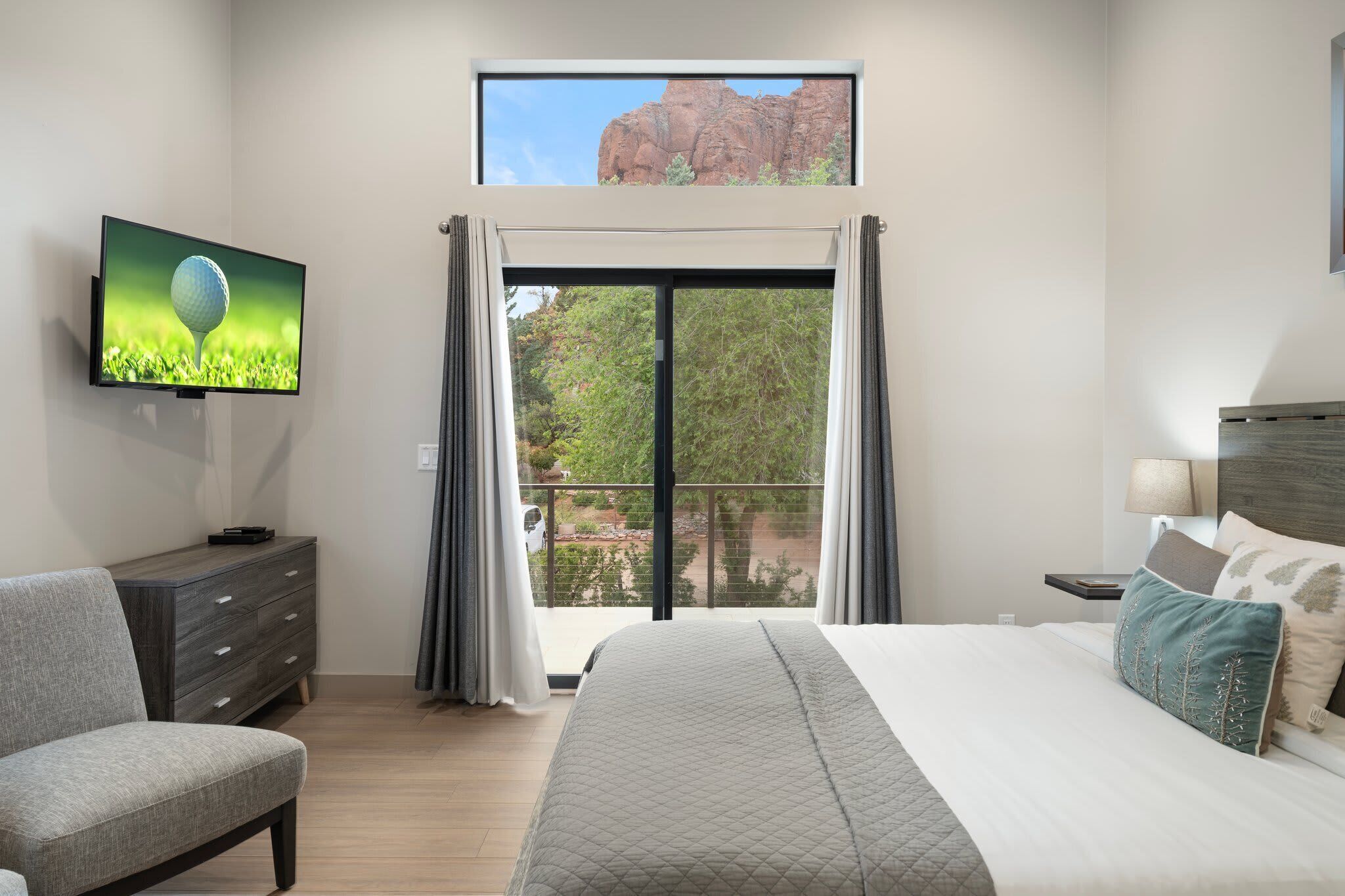 A bedroom with a bed, a TV, a dresser, and a balcony with a view of a rock formation.