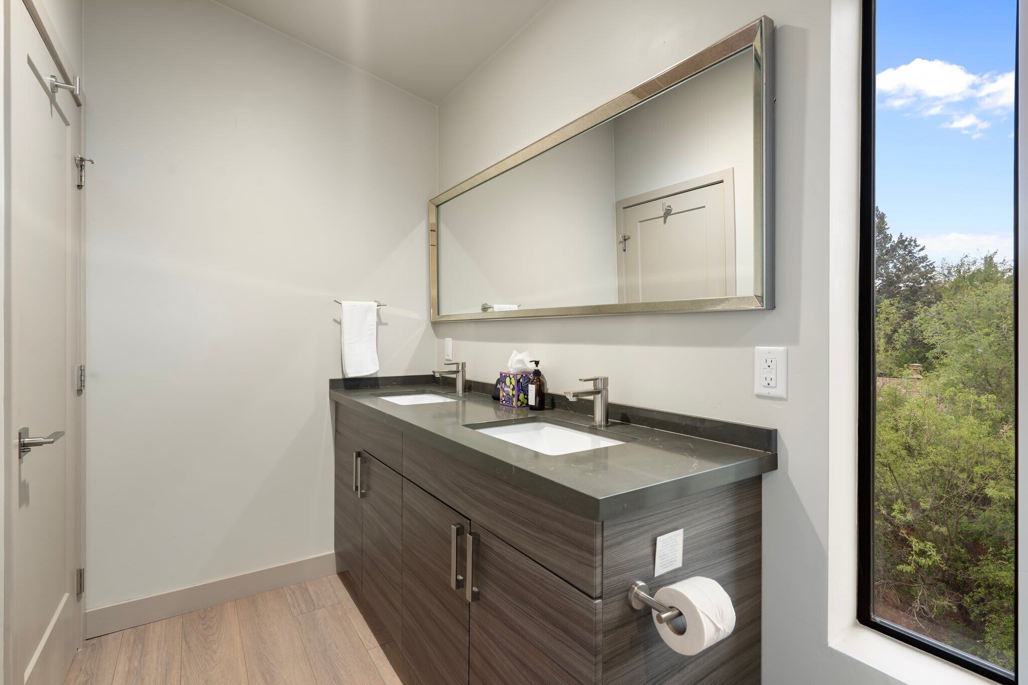 A bathroom with two sinks, a large mirror, and a window with a view of trees.