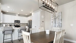 A modern kitchen with white cabinets, a stainless steel refrigerator, and a microwave. There is a dining area with a wooden table and white chairs. - NEW Alamo Alpine Retreat Sleeps 11 3 Showers (Flagstaff)