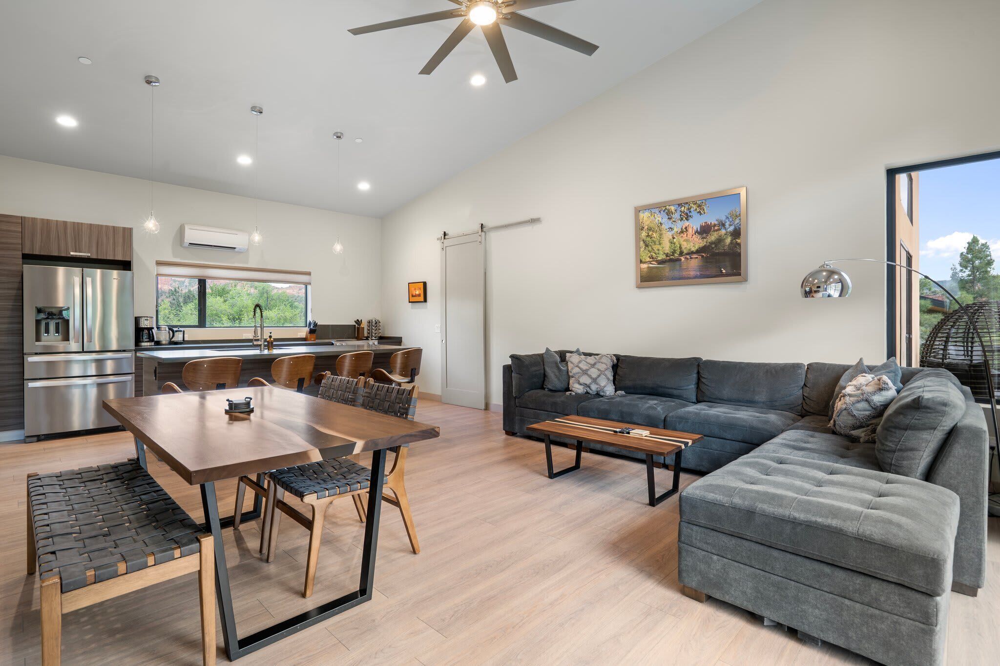 A modern living room with a grey sectional sofa, a wooden dining table with chairs, and a kitchen area with stainless steel appliances.