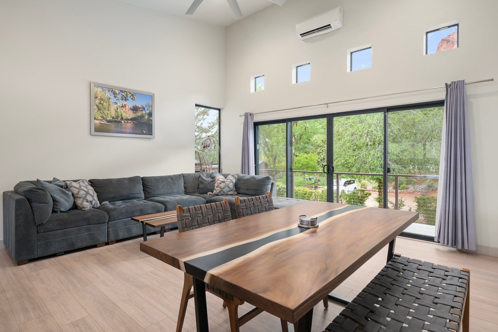 Una sala de estar moderna con un sofá gris, una mesa de comedor de madera y ventanas amplias que ofrecen vistas a zonas verdes y un coche.