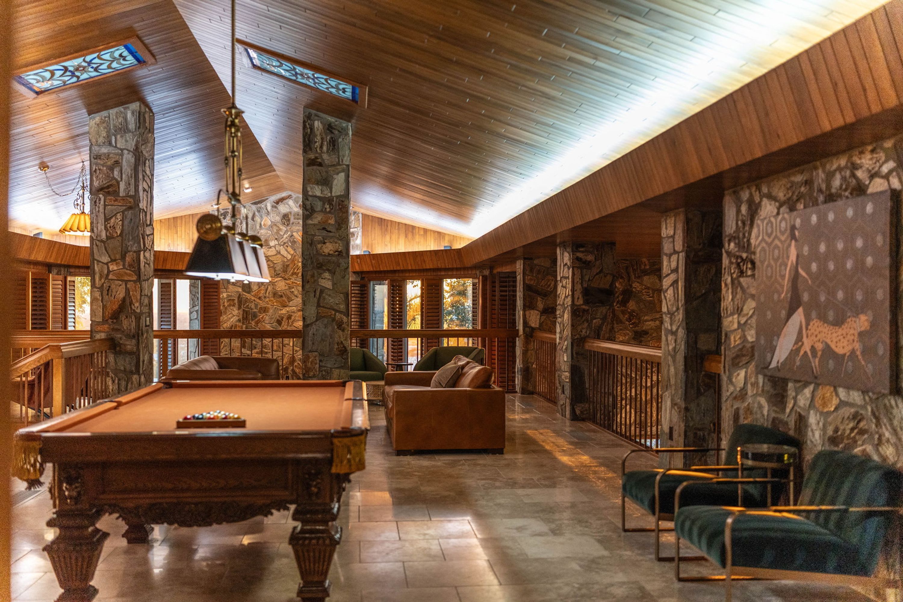 A pool table, a sofa, and a billiard cue rack in a room with stone walls and wooden ceilings.