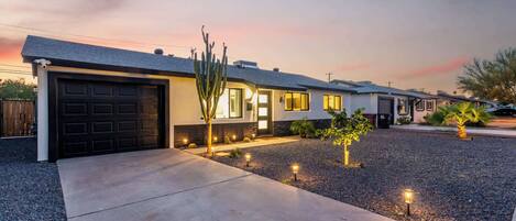 A modern two-story house with a dark garage door, a well-lit front yard with a cactus and small plants, and a clear sky at dusk.