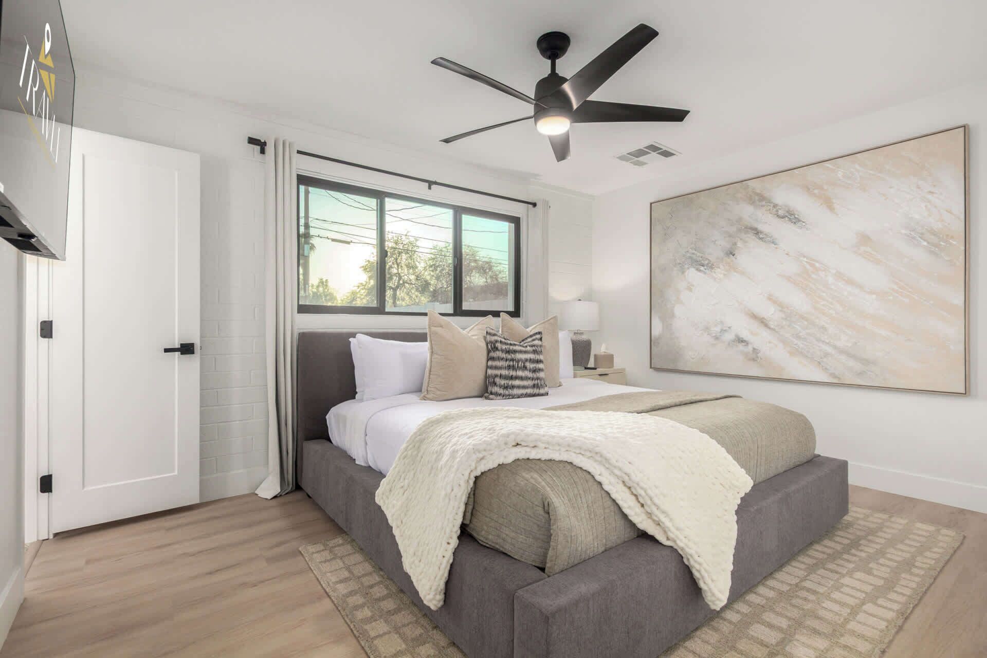 A bedroom with a bed, a ceiling fan, a window, and a large abstract painting on the wall.