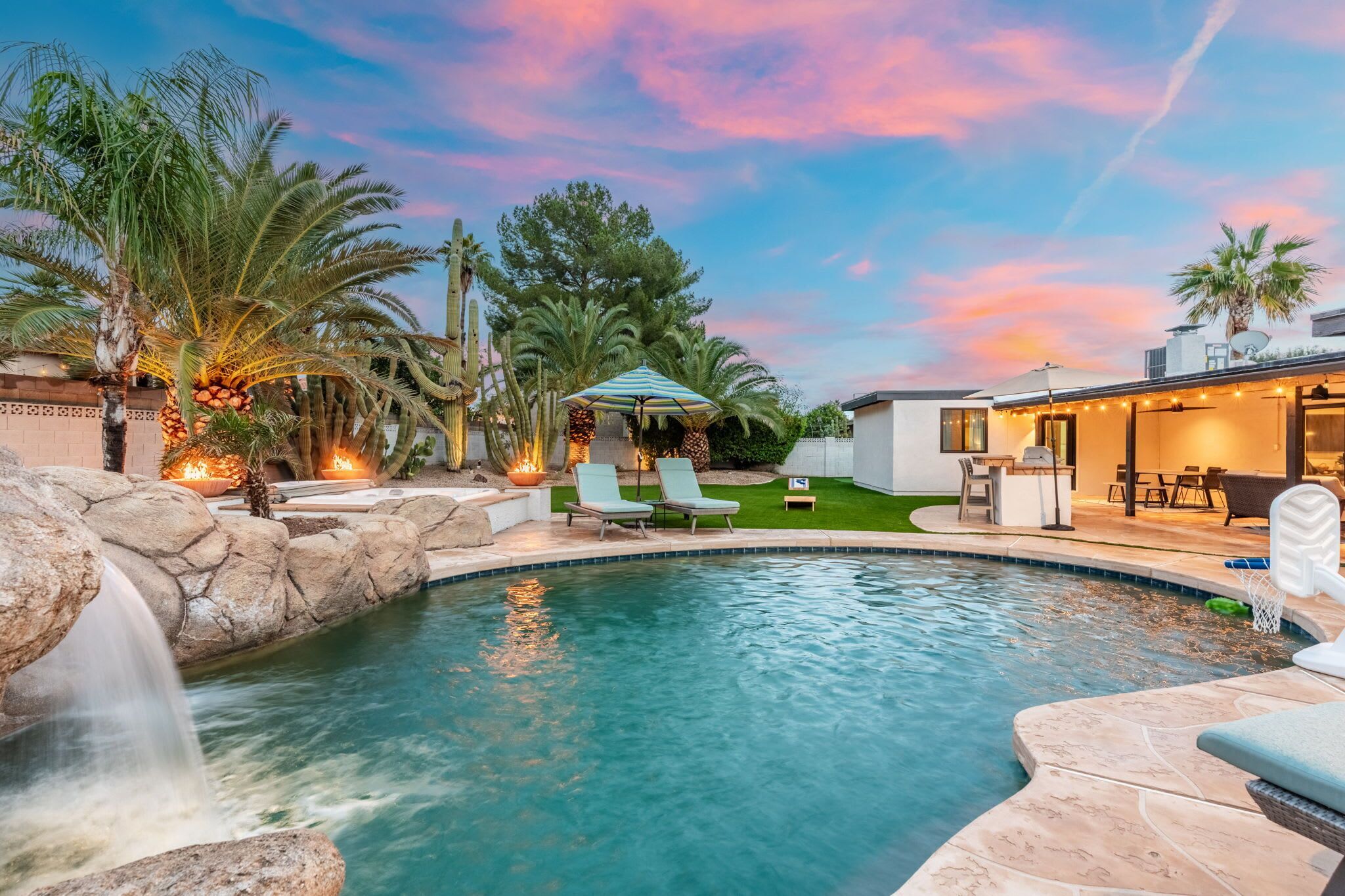 A swimming pool with a waterfall feature, surrounded by palm trees and a patio area with lounge chairs and a table.