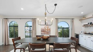 A dining area with a large round table, six chairs, a chandelier, and large windows with curtains. - NEW Grotto Falls Retreat Pool w Slide Spa Putt Grn (Scottsdale)