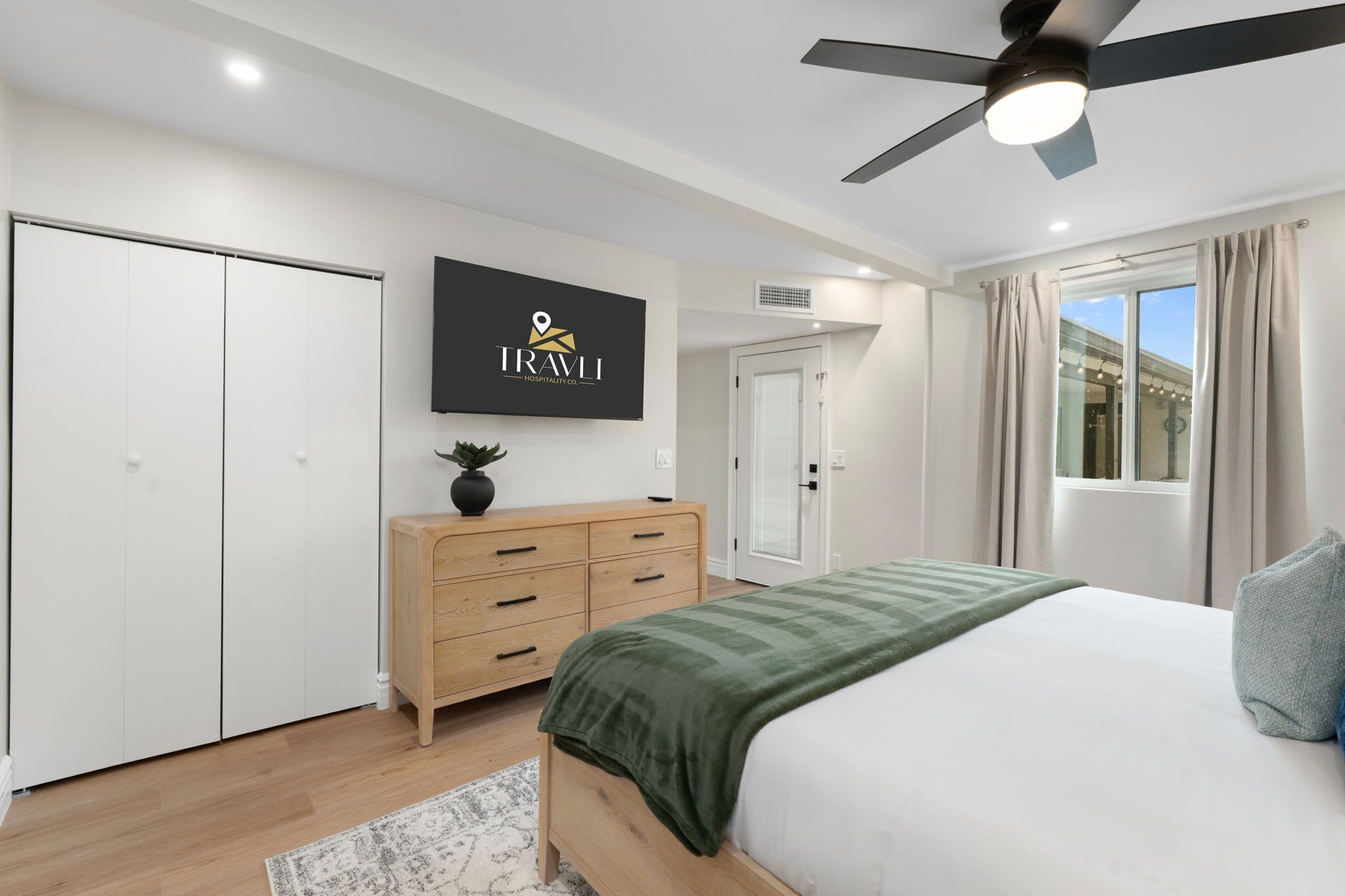 A modern hotel room with a bed, a wooden dresser, a flat-screen TV, a ceiling fan, and a view of the outside.