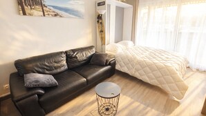 Et moderne soveværelse med en sort lædersofa, et rundt sofabord, en seng med en quiltet dyne, billedkunst med strandtema, en ventilator i loftet og lette gardiner.