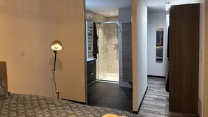 A modern hotel room with a bed, a bedside lamp, a bathroom with a shower, and a hallway leading to another room.